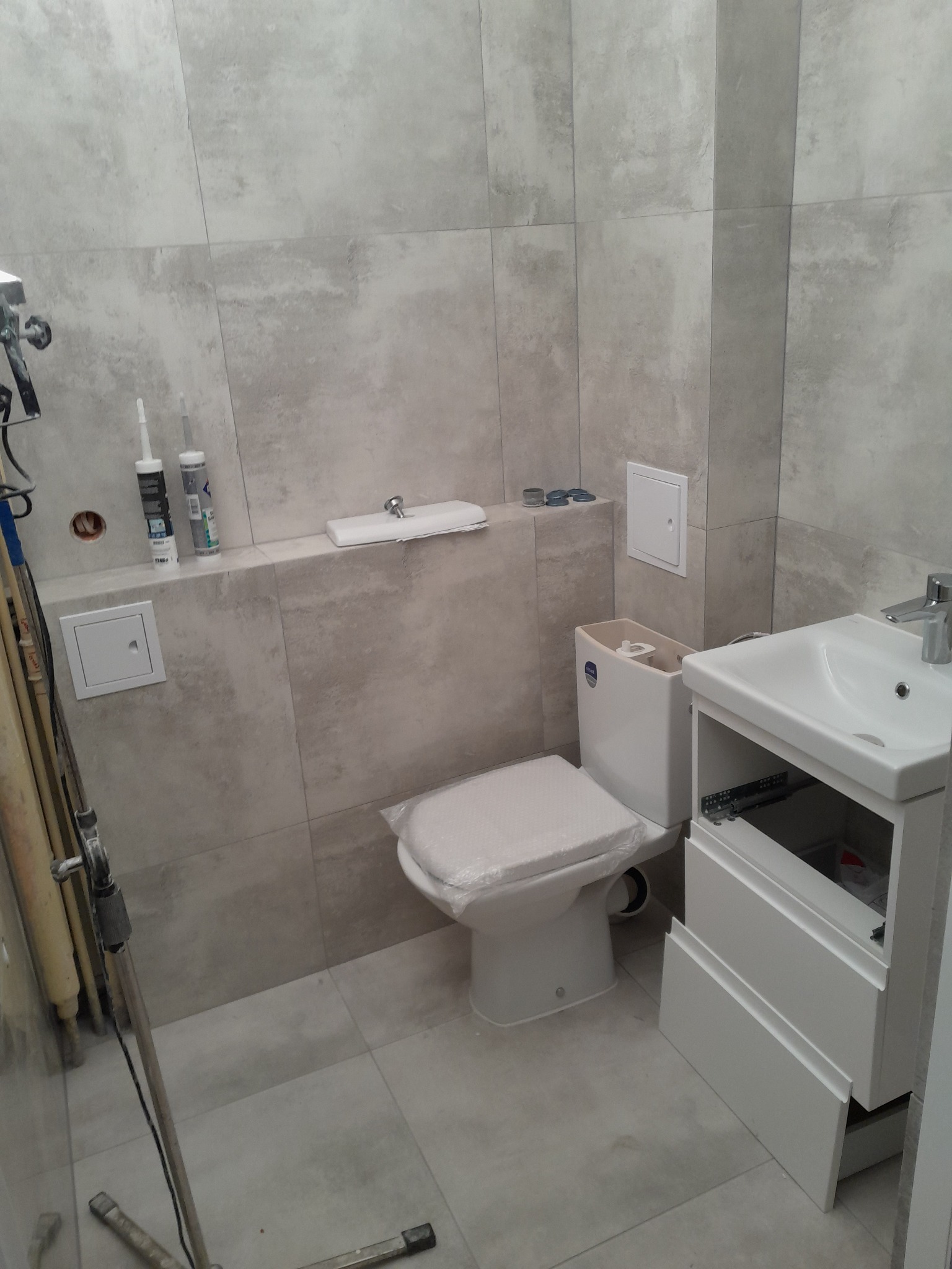 Jasna łazienka w trakcie wykańczania: płytki imitujące kamień, biała toaleta z deską w folii, umywalka z szafką bez frontów. Widoczne akcesoria i narzędzia.