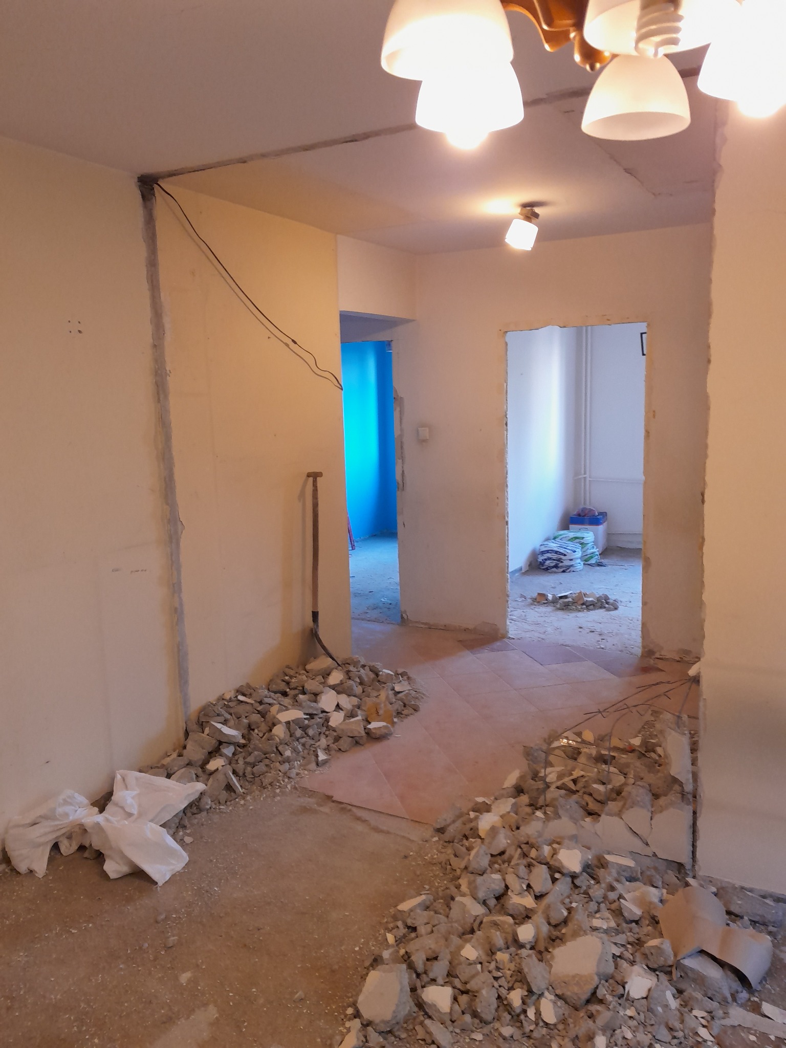 Wnętrze w trakcie demontażu: gruz, odsłonięte kable, ściany przygotowane do prac wykończeniowych. Szpadel oparty o ścianę. Widok na sąsiednie pomieszczenia.