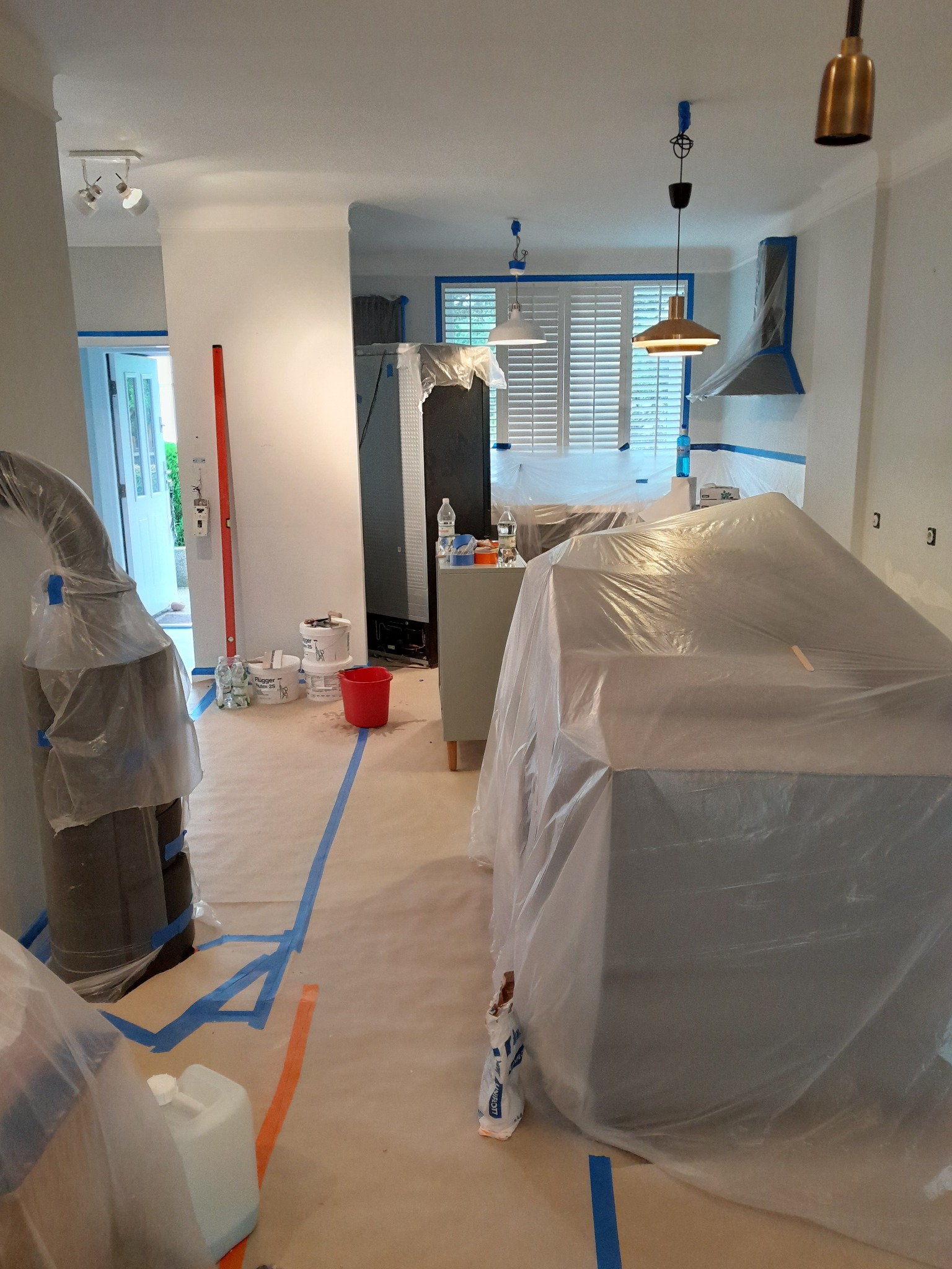 Przygotowania do malowania wnętrza: meble zabezpieczone folią, podłoga zakryta papierem, ściany oklejone taśmą malarską. Widoczne puszki z farbą i wiadro.