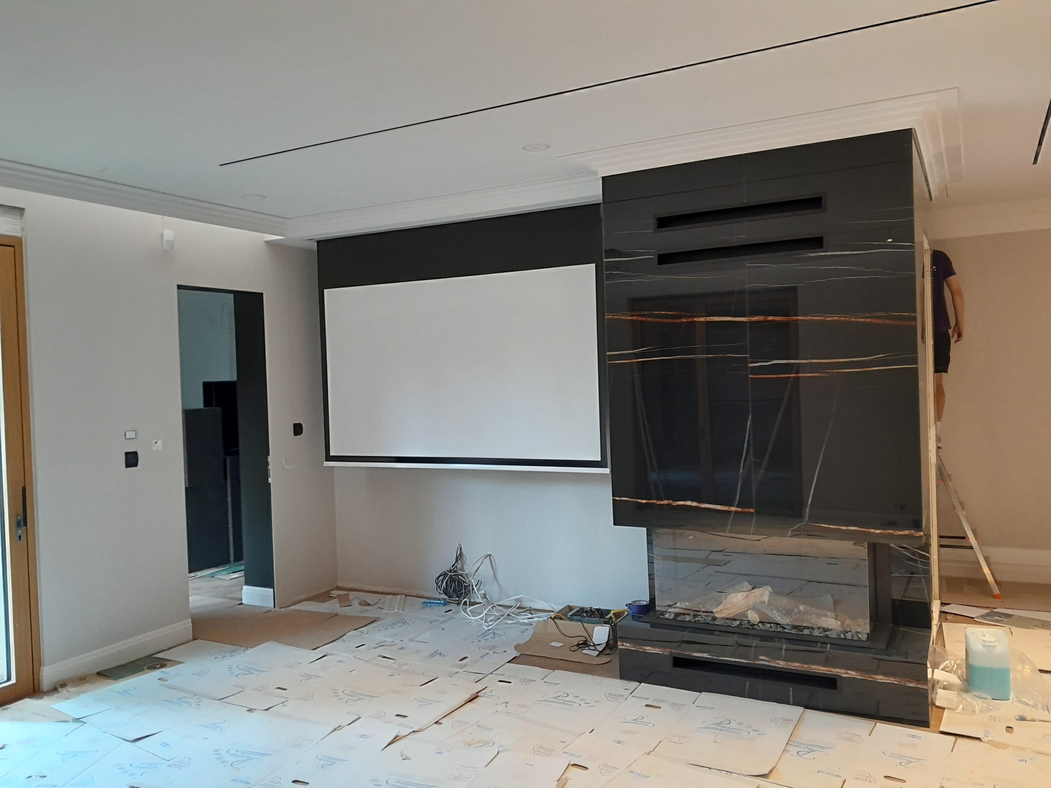 Wnętrze w trakcie wykańczania: marmurowy kominek z paleniskiem, ekran projekcyjny, podłoga zabezpieczona kartonem. Widoczny fragment drabiny i osoba na niej.