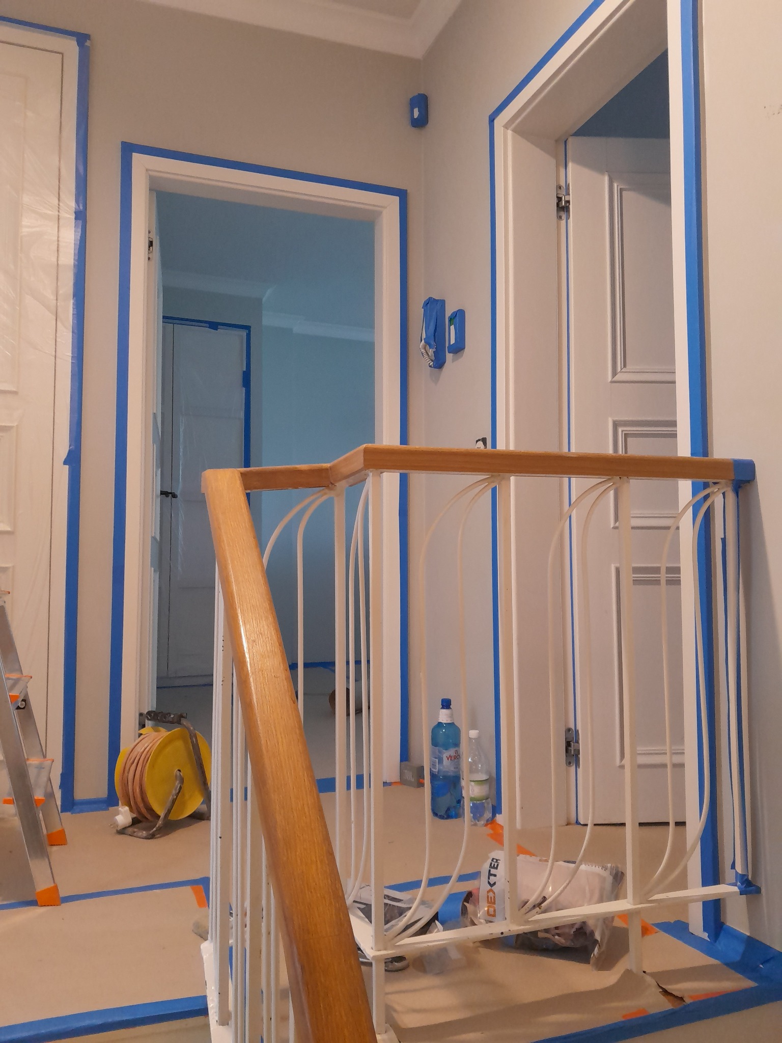 Korytarz w trakcie malowania: zabezpieczone taśmą malarską drzwi, ściany i schody. Widoczny fragment drabiny, butelki z wodą i narzędzia malarskie.
