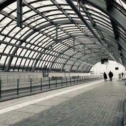 Artisso360 Aleksandra Biderman - Czarno-biała fotografia peronu dworcowego z zadaszeniem łukowym, gdzie w oddali widać idących ludzi. Minimalistyczny kadr z perspektywą głębi.
