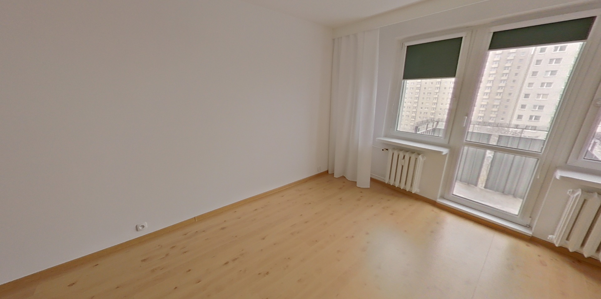 Pusty pokój z drewnianą podłogą, białymi ścianami i dużymi oknami z widokiem na budynek mieszkalny. Grzejniki pod oknami. Przygotowanie do wirtualnego meblowania.