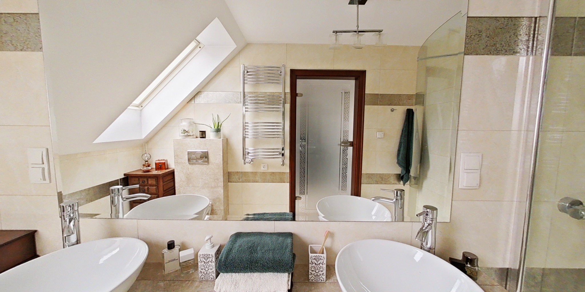 Jasna łazienka z dwoma umywalkami w stylu wolnostojącym, lustrem na całą ścianę i oknem dachowym. Widoczny grzejnik drabinkowy i kabina prysznicowa.