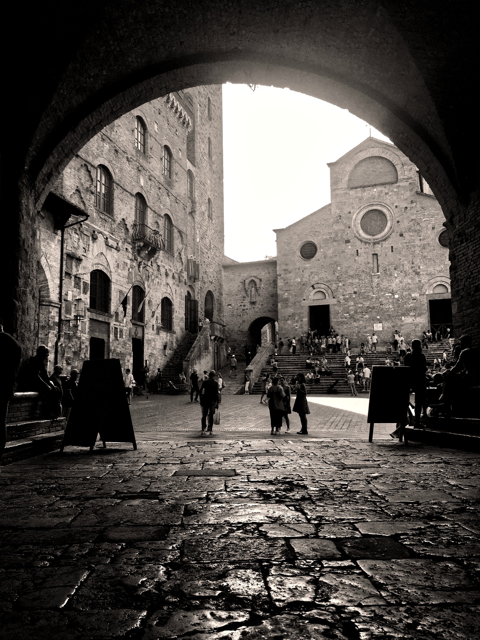 Czarno-biała fotografia ukazująca kamienną uliczkę, budynki i kościół w San Gimignano, widziane przez kamienny łuk. Ludzie w tle dodają życia scenerii.