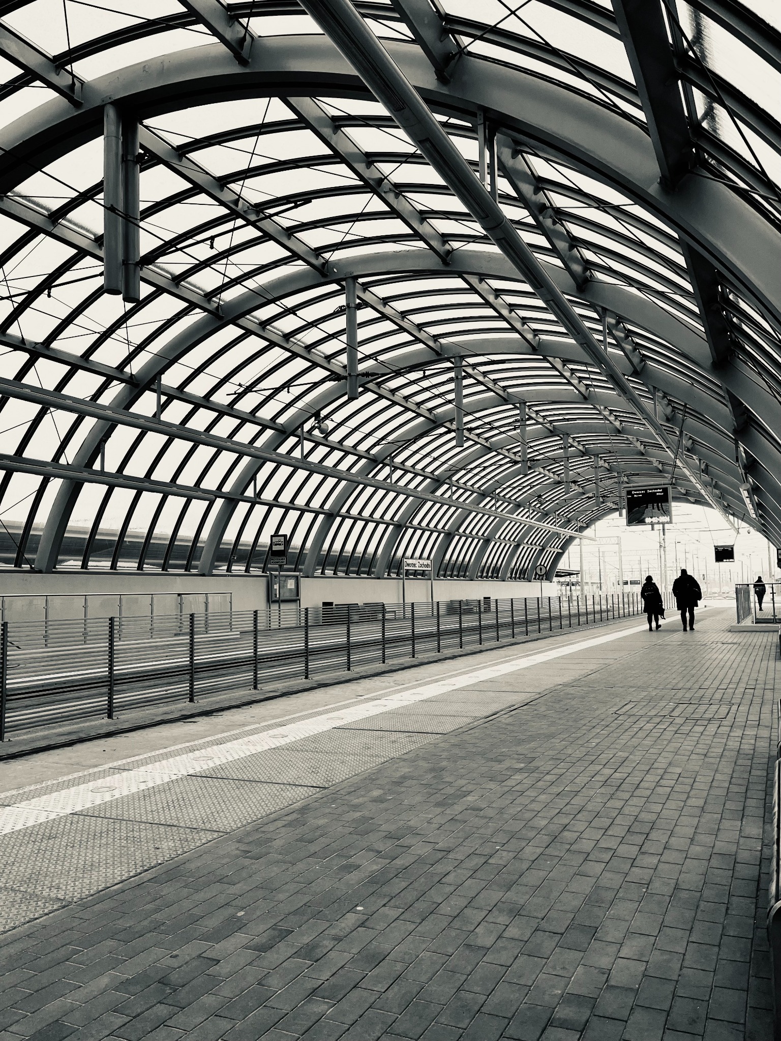 Czarno-biała fotografia peronu dworcowego z zadaszeniem łukowym, gdzie w oddali widać idących ludzi. Minimalistyczny kadr z perspektywą głębi.