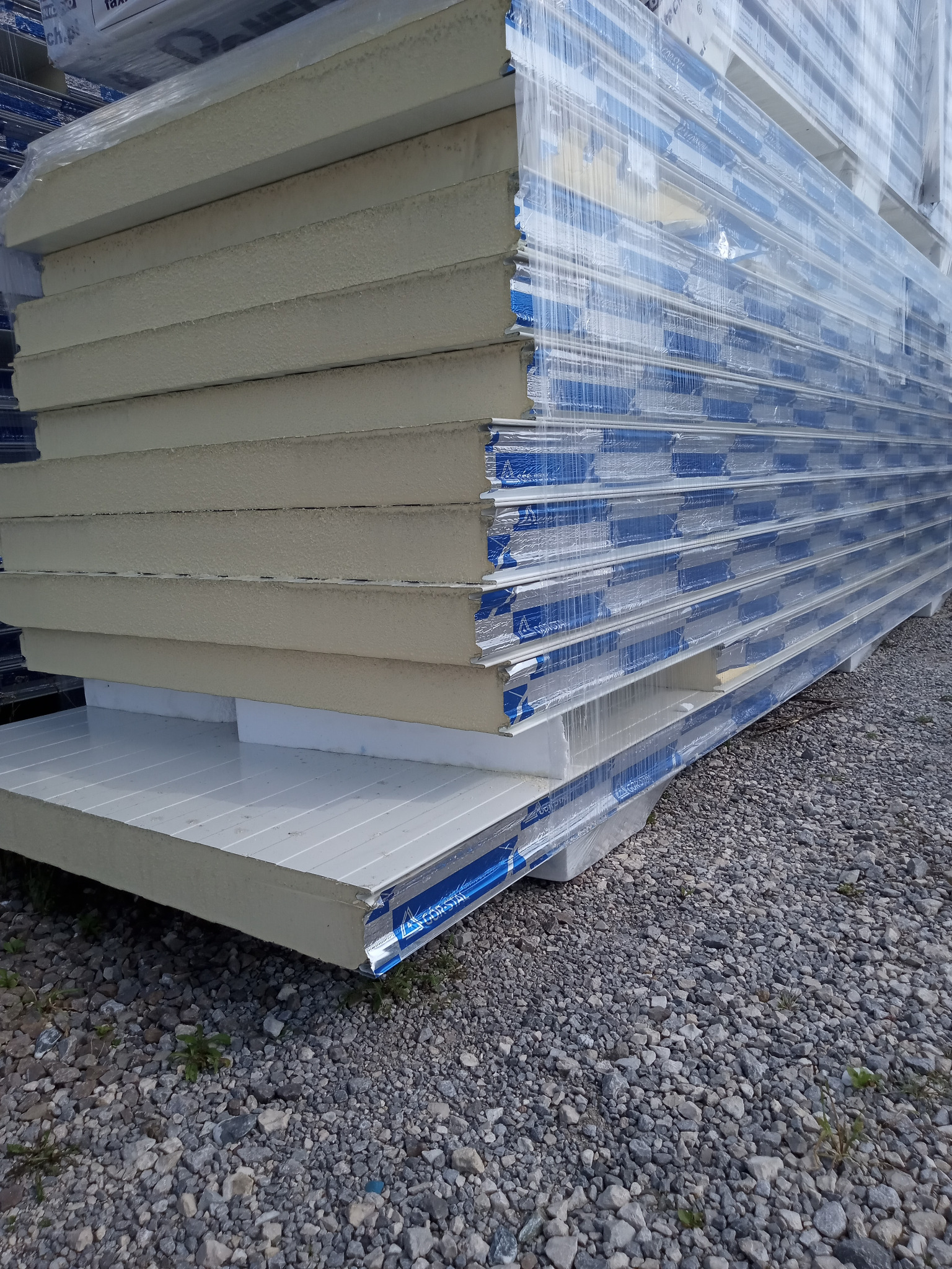 Stos płyt warstwowych PIR (biały/linia) o grubości 10cm, owiniętych folią, leżących na żwirowym podłożu. Widoczny detal krawędzi płyty z niebieskim oznaczeniem.