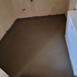 BEESYSTEM SPÓŁKA Z OGRANICZONĄ ODPOWIEDZIALNOŚCIĄ - Świeżo wylana, gładka posadzka jastrychowa w pomieszczeniu z widoczną skrzynką instalacyjną przy ścianie. Szary odcień wylewki, surowe ściany, widoczne gniazdka.