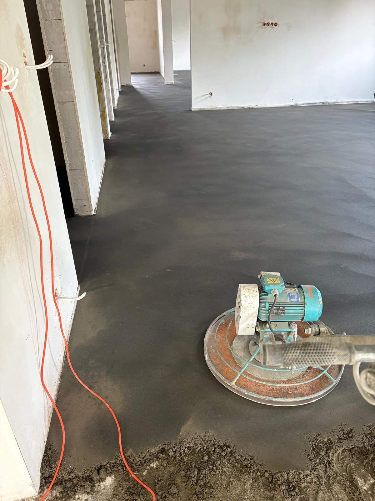 Wyrównana, szara posadzka w surowym wnętrzu, z maszyną do wygładzania i wystającymi kablami. Widok na długie, puste pomieszczenie w stanie deweloperskim.