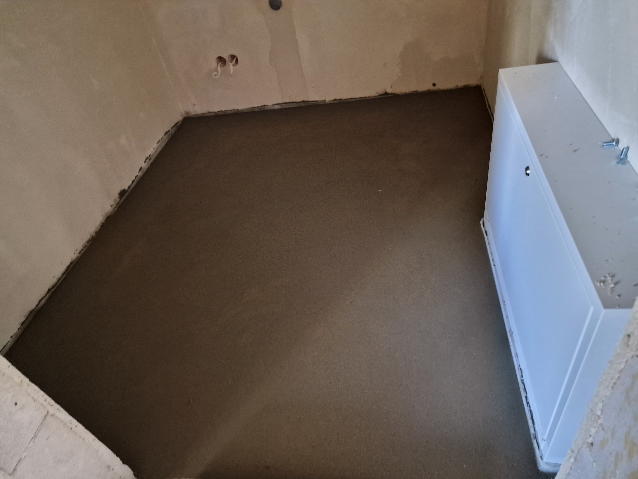 Świeżo wylana, gładka posadzka jastrychowa w pomieszczeniu z widoczną skrzynką instalacyjną przy ścianie. Szary odcień wylewki, surowe ściany, widoczne gniazdka.