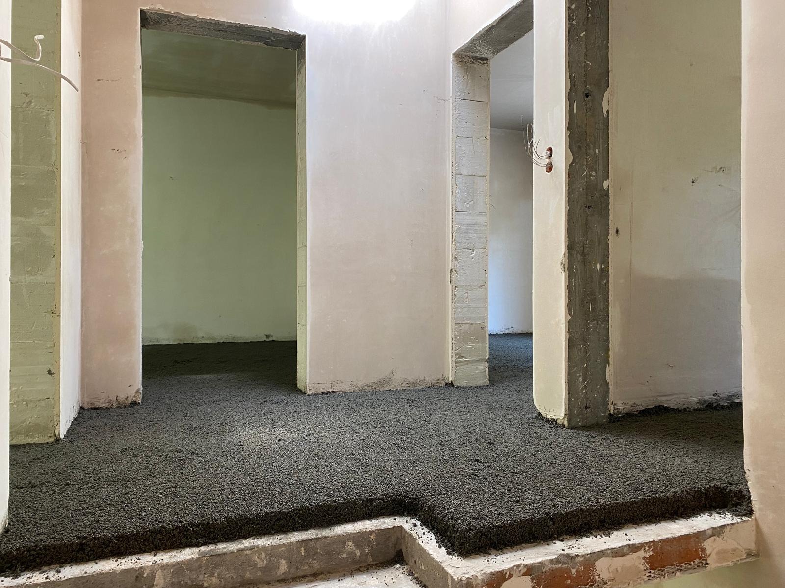 Wnętrze w trakcie prac: świeżo wylana, ciemna posadzka jastrychowa w pomieszczeniu z surowymi ścianami i otworami drzwiowymi. Widoczny etap prac wykończeniowych.