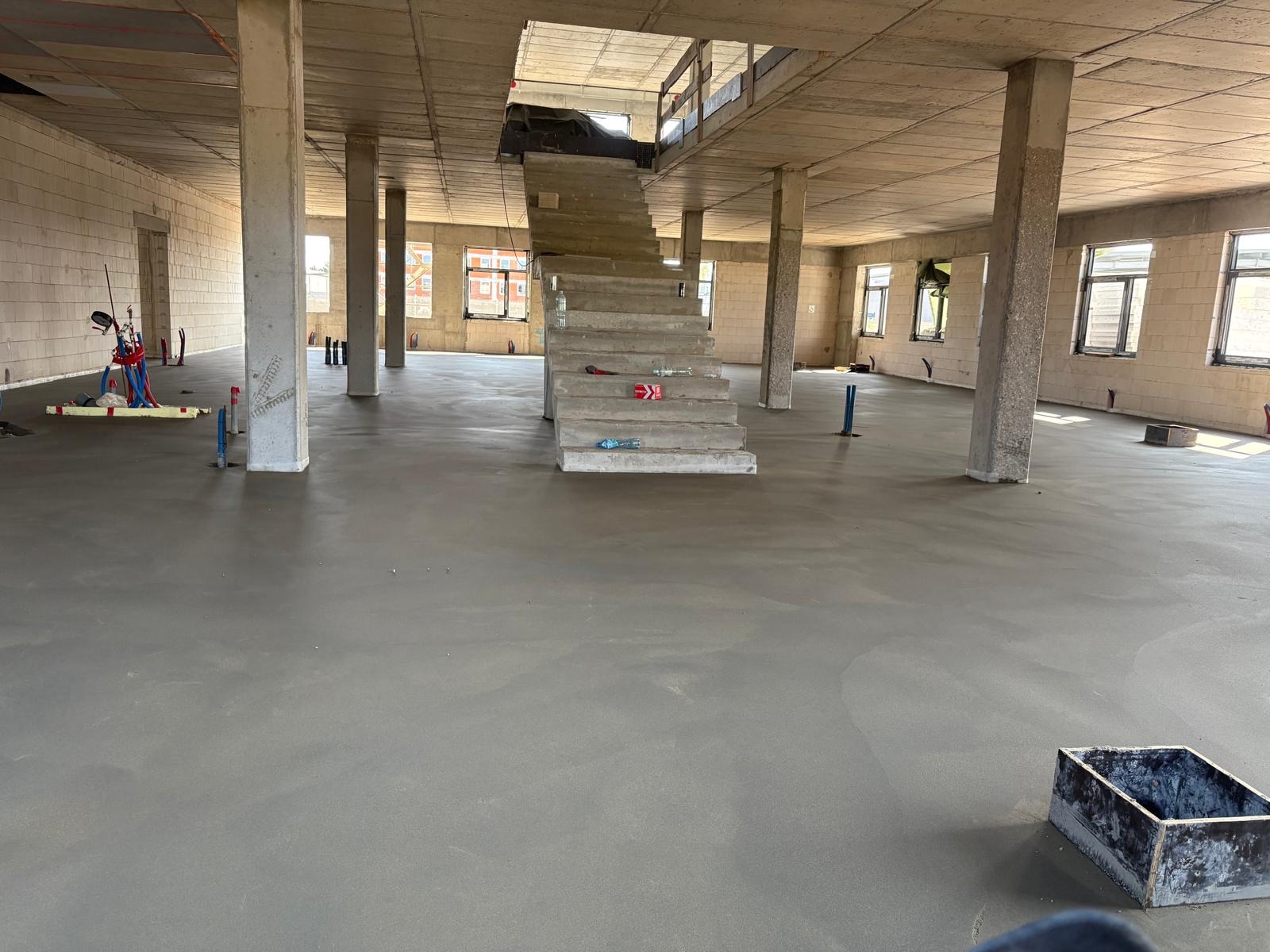 Wnętrze w stanie surowym z wylaną, gładką posadzką, betonowymi kolumnami i schodami. Widoczne okna, narzędzia i akcesoria budowlane.
