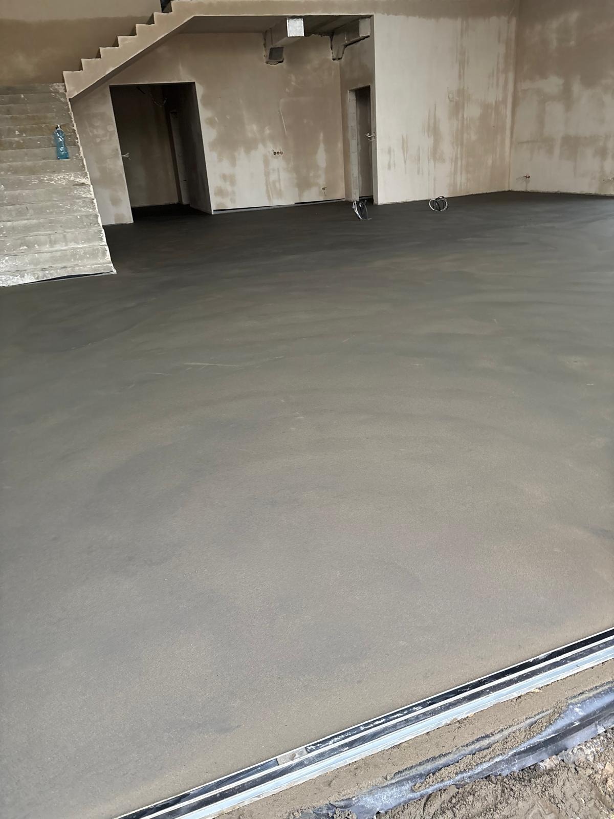 Świeżo wylana, gładka posadzka w surowym wnętrzu z widocznymi schodami i otworami drzwiowymi. Minimalistyczny wystrój, naturalne światło.