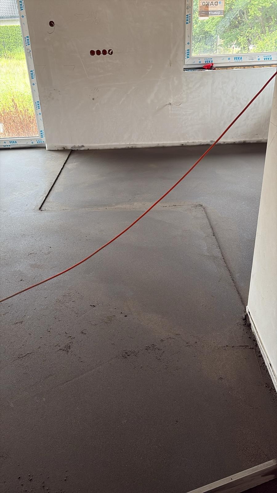 Świeżo wylana, szara posadzka jastrychowa w surowym wnętrzu z oknem. Widoczne dylatacje i czerwony kabel na podłodze. Ściany przygotowane do dalszych prac wykończeniowych.
