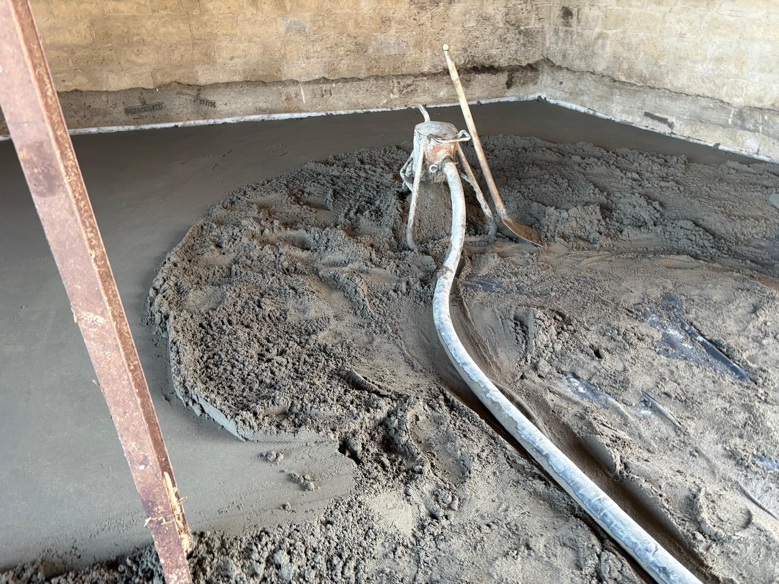 Wylewka posadzki z użyciem węża i mieszadła. Świeża, szara wylewka pokrywa podłoże, widoczne narzędzia pracy. Surowy stan, przygotowanie do dalszych prac wykończeniowych.