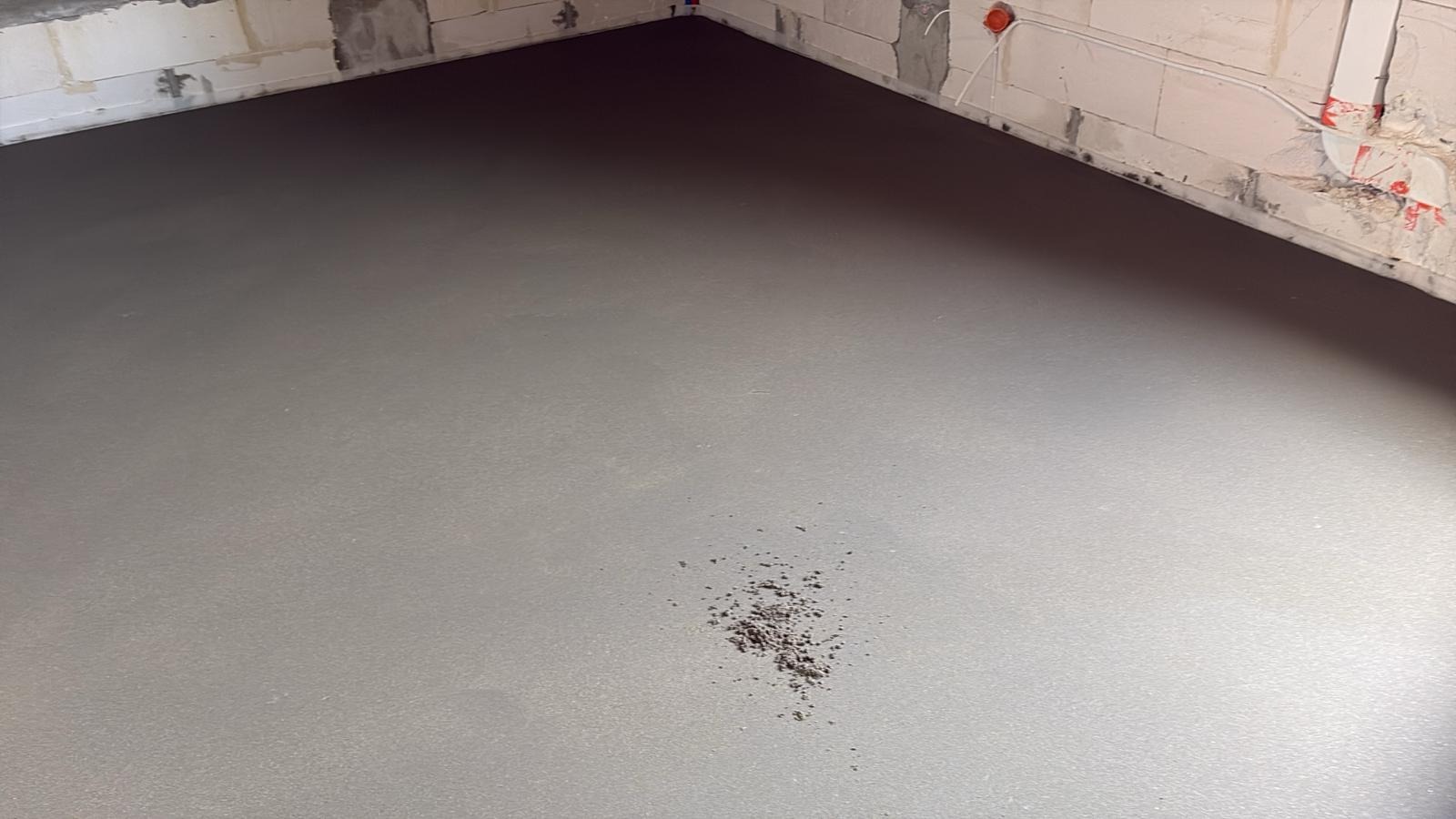 Gładka, szara wylewka w surowym wnętrzu z widocznymi bloczkami i instalacją elektryczną. Na powierzchni wylewki widoczne drobne zabrudzenia.