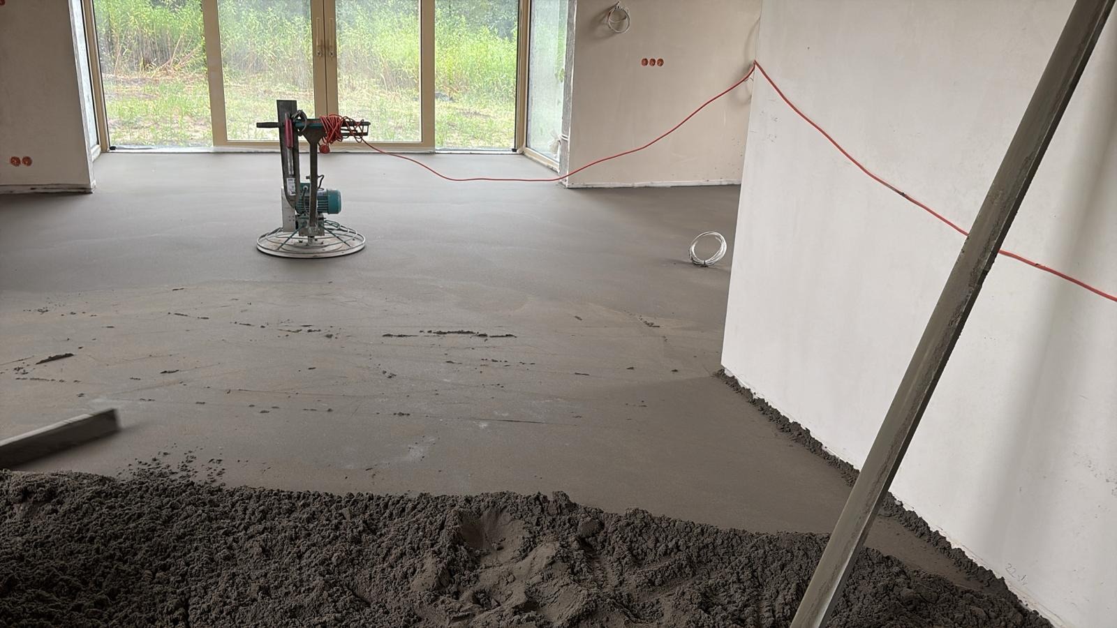 Wyrównywanie wylewki jastrychowej w jasnym wnętrzu z dużymi oknami. Widoczna maszyna do zacierania i narzędzia. Świeża, szara wylewka na podłodze.