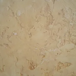 Beżowa ściana pokryta tynkiem strukturalnym o nieregularnej, chropowatej fakturze z widocznymi zagłębieniami i wypukłościami, imitująca stary tynk.