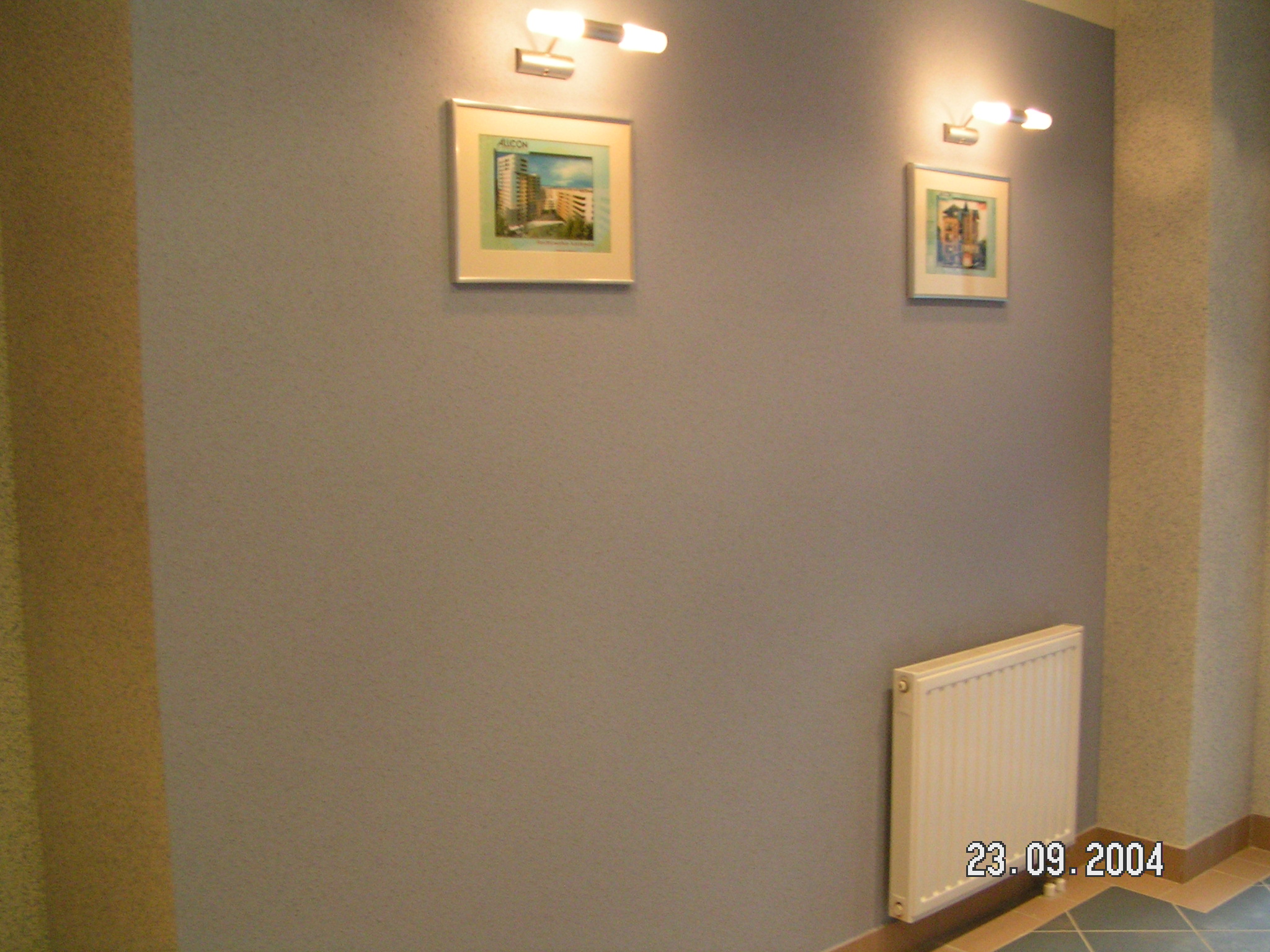 Ściana pomalowana na szaro, z fakturą przypominającą natryskową, oświetlona dwoma kinkietami z żarówkami halogenowymi, z dwoma obrazkami w ramkach i grzejnikiem panelowym u dołu.