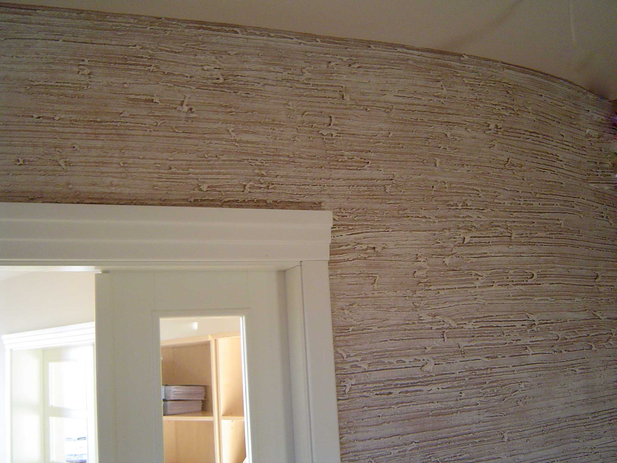 Beżowa ściana z tynkiem strukturalnym o wyraźnej, poziomej fakturze, widoczna z perspektywy wnętrza, obok białej futryny drzwi.