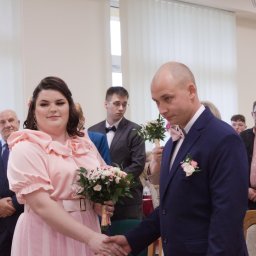 Daria Śmiechowska - Panna młoda w pudrowej sukni i pan młody w granatowym garniturze ślubnym ściskają sobie dłonie podczas ceremonii, w tle goście i jasne wnętrze.