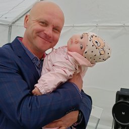 Daria Śmiechowska - Uśmiechnięty mężczyzna w garniturze trzyma na rękach małą dziewczynkę w czapce z motywem zwierzęcym, w tle biały namiot i elementy nagłośnienia.