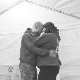 Daria Śmiechowska - Czarno-białe zdjęcie pary w czułym uścisku pod białym namiotem, z widocznymi plastikowymi skrzynkami w tle. Mężczyzna w bluzie i spodniach moro, kobieta w ciemnych legginsach.