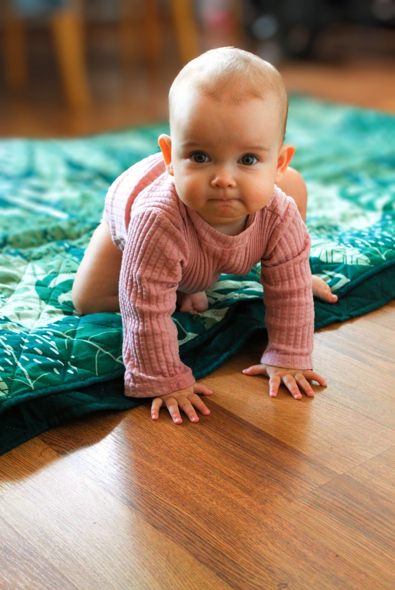 Urocze, pełzające niemowlę w różowym ubranku na zielonej, wzorzystej macie, na tle parkietu. Dziecko patrzy wprost w obiektyw, wyraz twarzy skupiony.