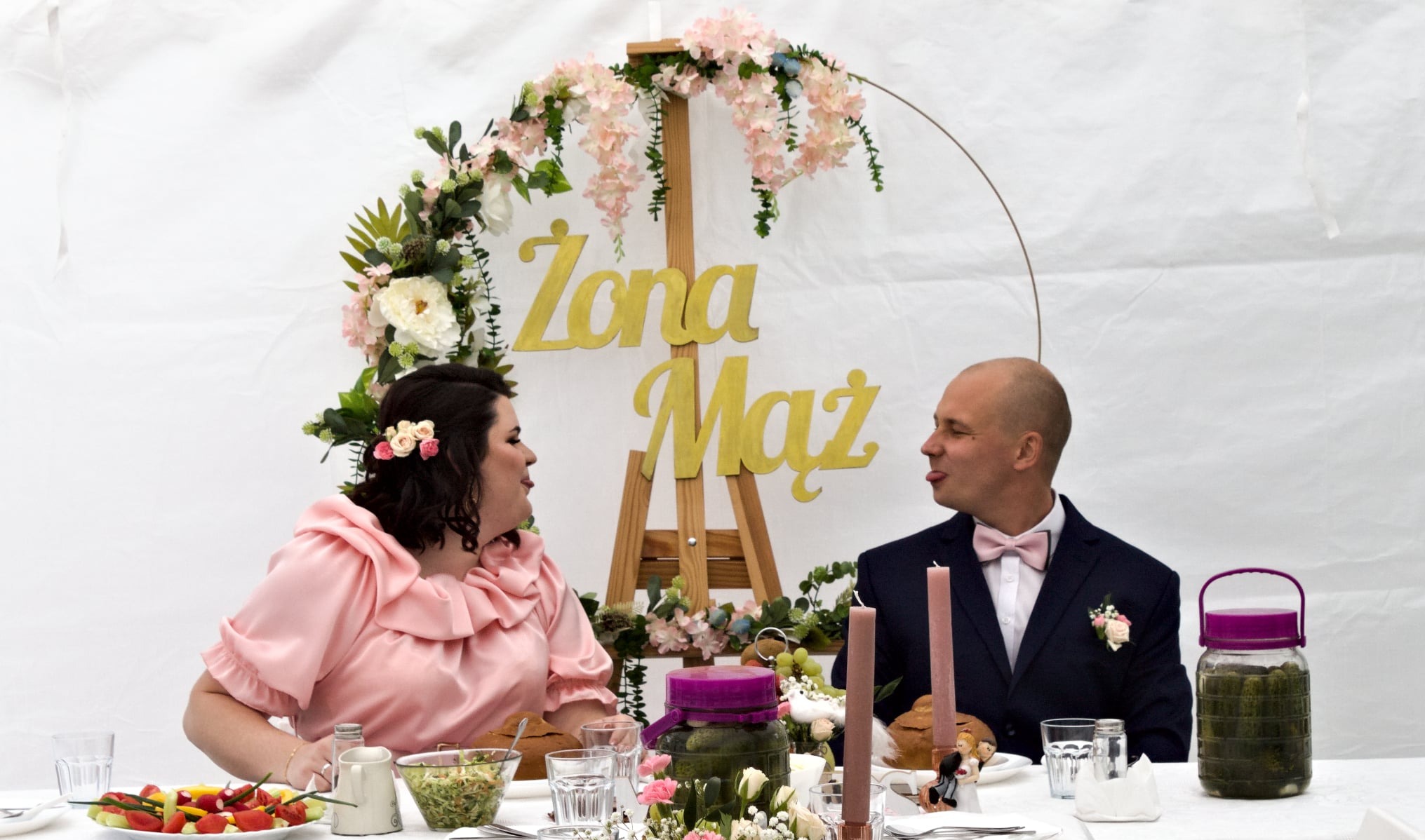 Para młoda siedzi przy stole weselnym z dekoracją 'Żona Mąż', mężczyzna pokazuje język. Na stole jedzenie i słoiki z ogórkami.