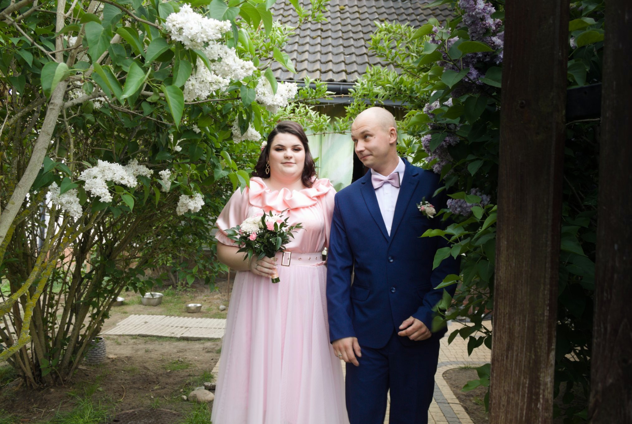 Uśmiechnięta para w strojach wizytowych pozuje wśród kwitnących bzów: ona w pudrowej sukni z bukietem, on w granatowym garniturze z muszką.