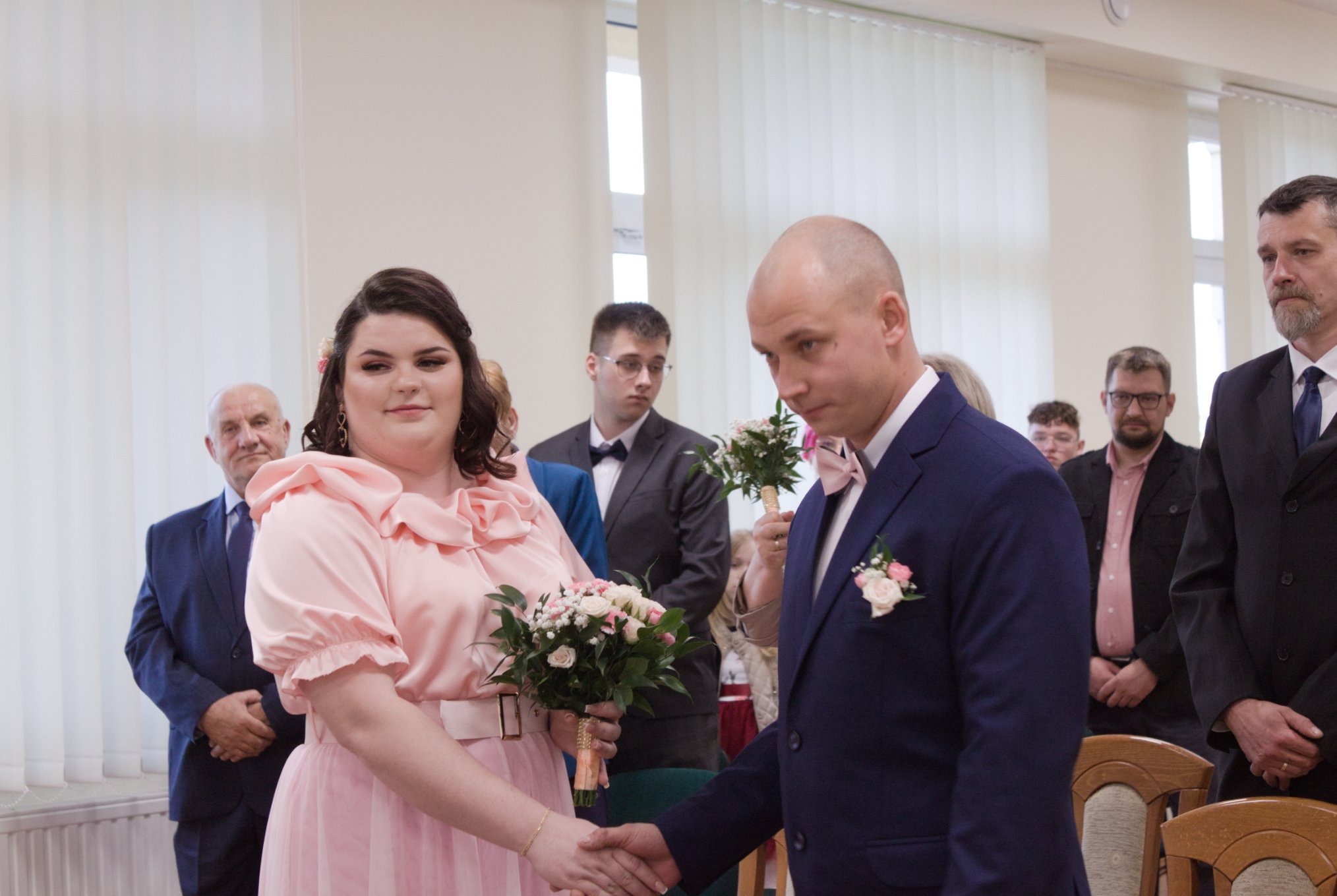 Panna młoda w pudrowej sukni i pan młody w granatowym garniturze ślubnym ściskają sobie dłonie podczas ceremonii, w tle goście i jasne wnętrze.