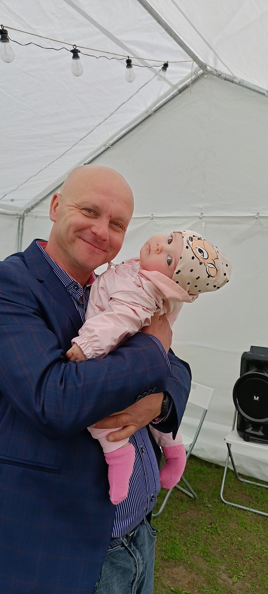 Uśmiechnięty mężczyzna w garniturze trzyma na rękach małą dziewczynkę w czapce z motywem zwierzęcym, w tle biały namiot i elementy nagłośnienia.