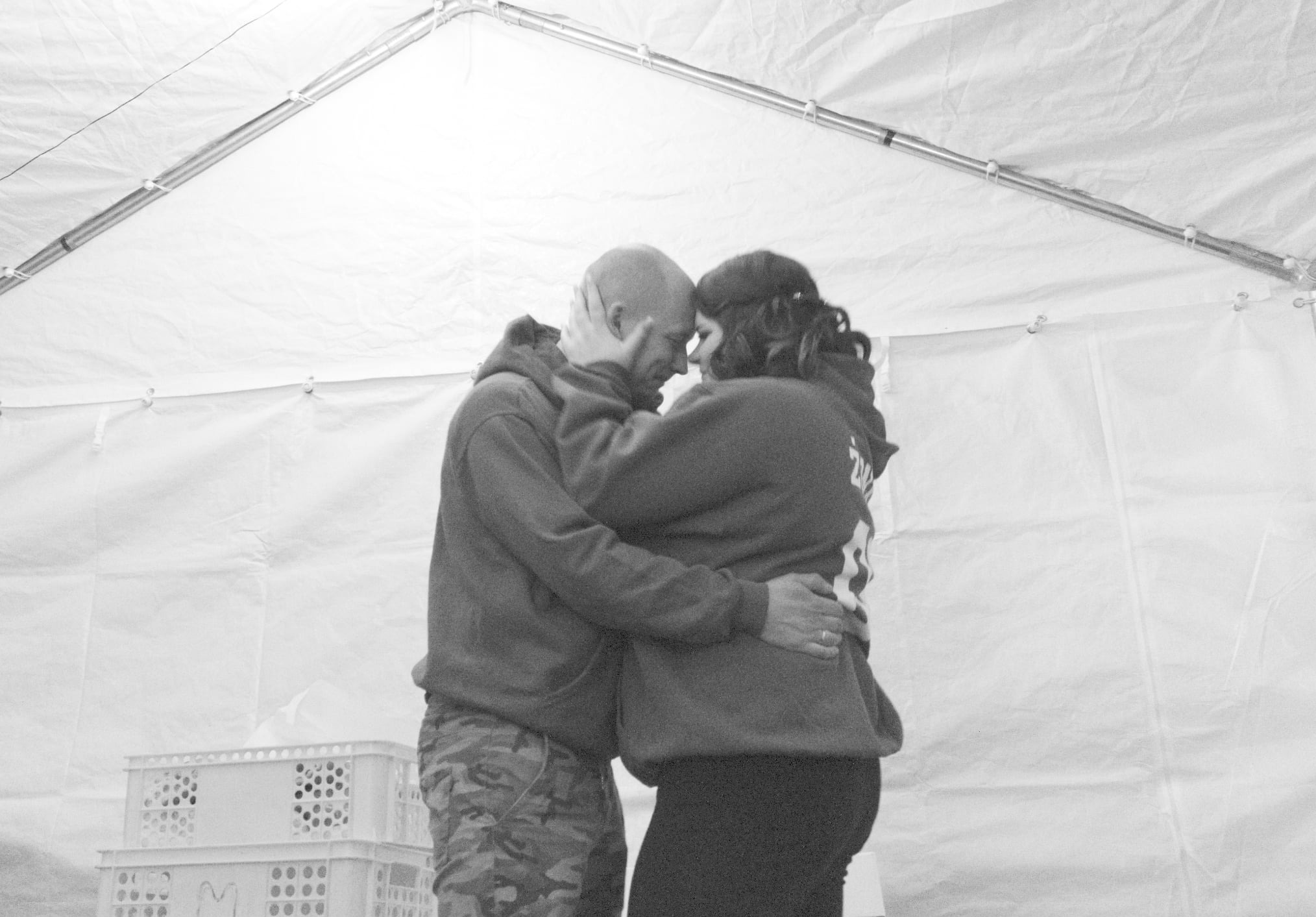 Czarno-białe zdjęcie pary w czułym uścisku pod białym namiotem, z widocznymi plastikowymi skrzynkami w tle. Mężczyzna w bluzie i spodniach moro, kobieta w ciemnych legginsach.