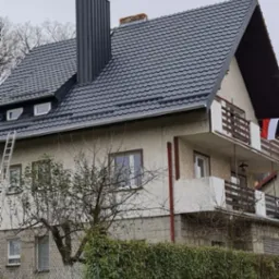 Dom z nowym, grafitowym dachem z blachodachówki i wysokim kominem. Przy ścianie oparta drabina. Ujęcie z poziomu ulicy, w tle drzewa bez liści.