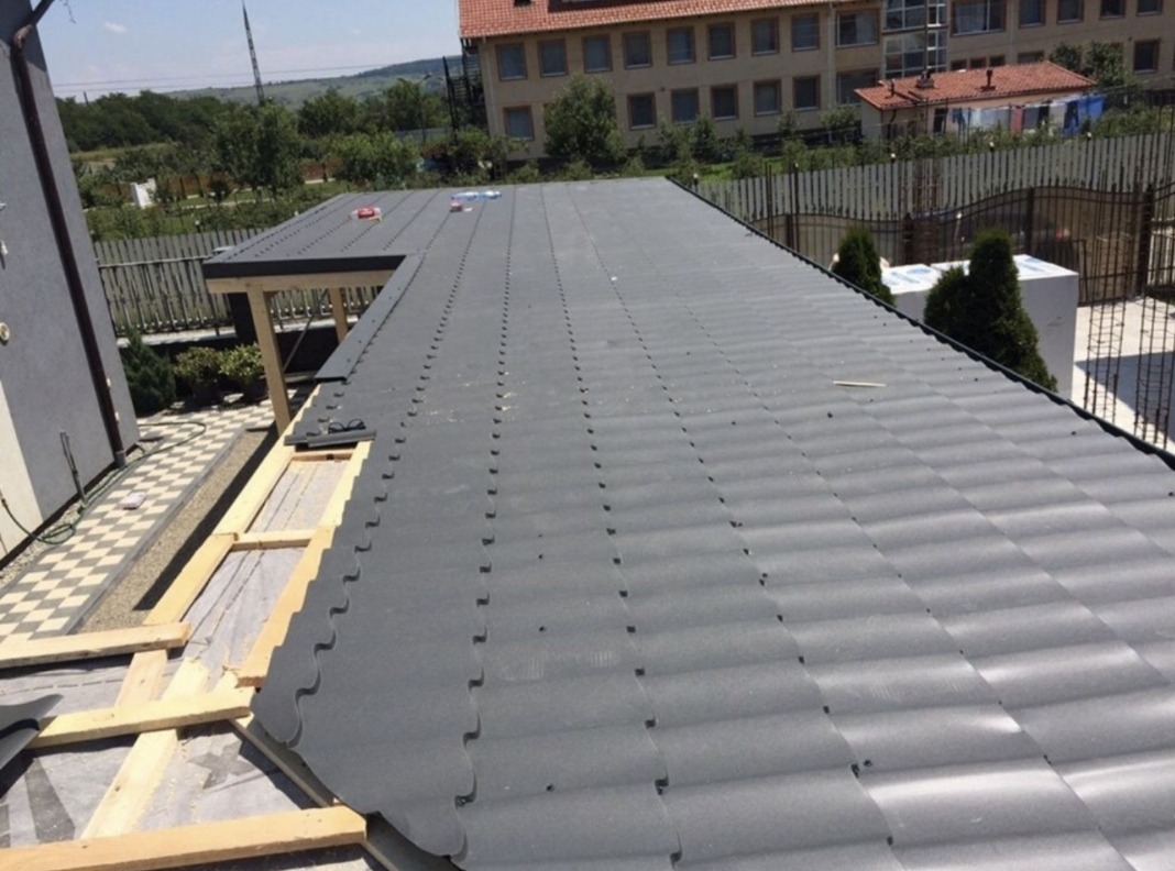 Częściowy widok szarego dachu z blachy trapezowej w trakcie montażu. Widoczne drewniane elementy konstrukcji, tło stanowi budynek mieszkalny i zieleń.