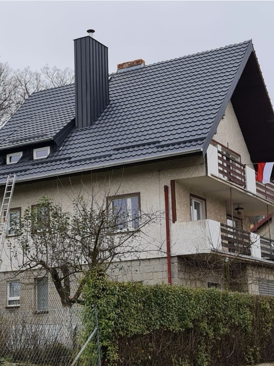 Dom z nowym, grafitowym dachem z blachodachówki i wysokim kominem. Przy ścianie oparta drabina. Ujęcie z poziomu ulicy, w tle drzewa bez liści.