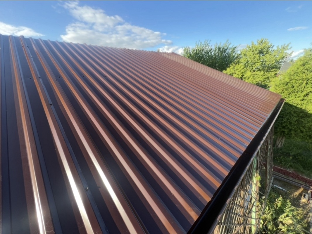 Nowy, brązowy dach z blachy trapezowej w promieniach słońca. Widok z góry ukazuje regularne rowki i odbicia światła. W tle zieleń drzew i błękit nieba.