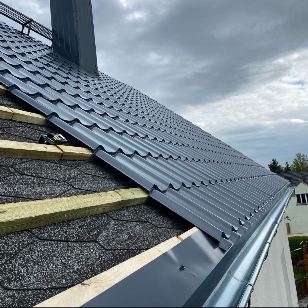 Montaż grafitowej blachy trapezowej na dachu z papy bitumicznej, widoczne łaty drewniane i rynna w kolorze blachy. Pochmurne niebo w tle.