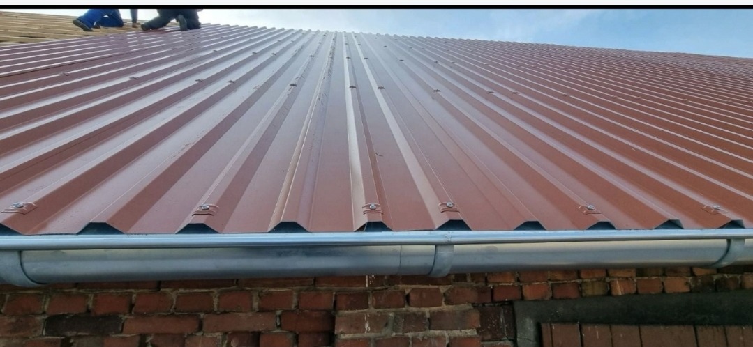 Nowe, brązowe krycie dachu z blachy trapezowej, zamontowane rynny, widoczny fragment drewnianej więźby i muru z czerwonej cegły. Ujęcie z dołu.