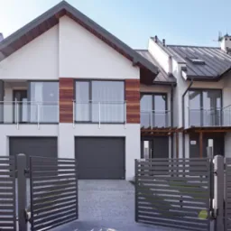 Nowoczesny dom bliźniak z szarą, automatyczną bramą wjazdową Wiśniowski. Elewacja biała z drewnianymi akcentami. Balkony z szklanymi balustradami. Słoneczny dzień.