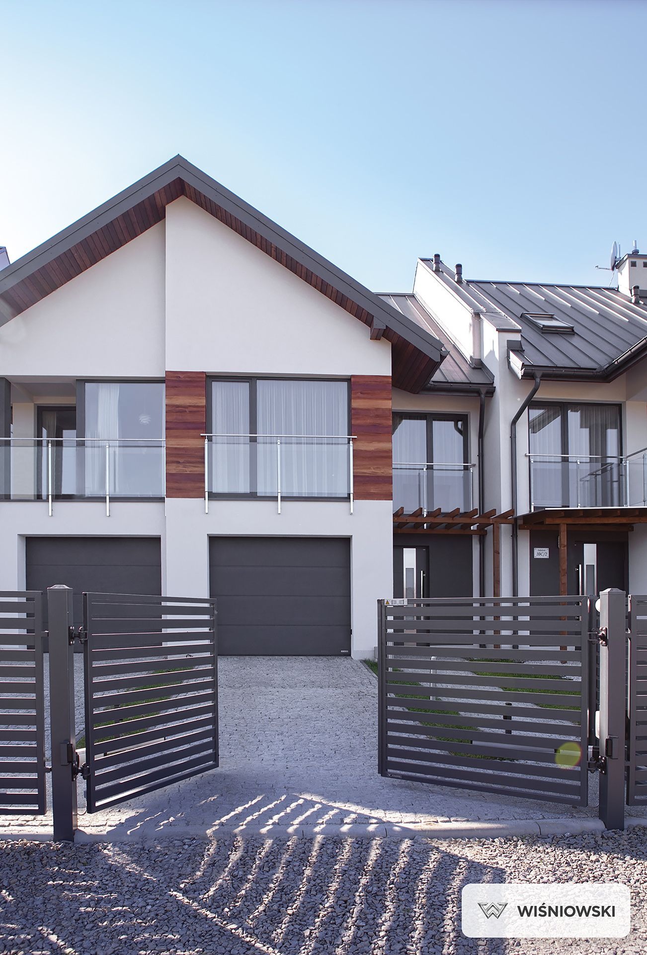 Nowoczesny dom bliźniak z szarą, automatyczną bramą wjazdową Wiśniowski. Elewacja biała z drewnianymi akcentami. Balkony z szklanymi balustradami. Słoneczny dzień.