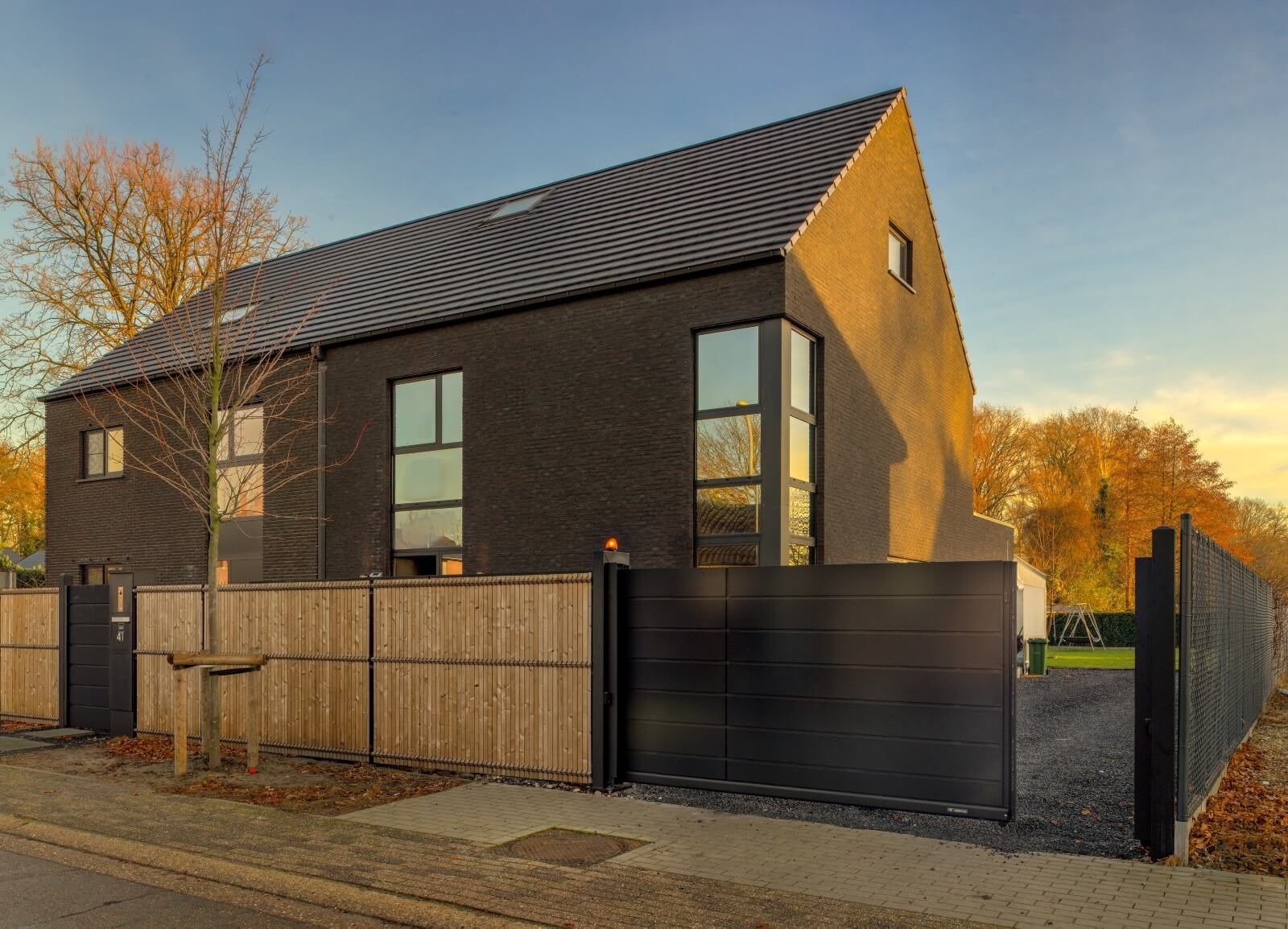 Nowoczesny dom z czarnej cegły z dużymi oknami. Brama wjazdowa przesuwna, metalowa, w kolorze antracytowym. Częściowo widoczne drewniane ogrodzenie.