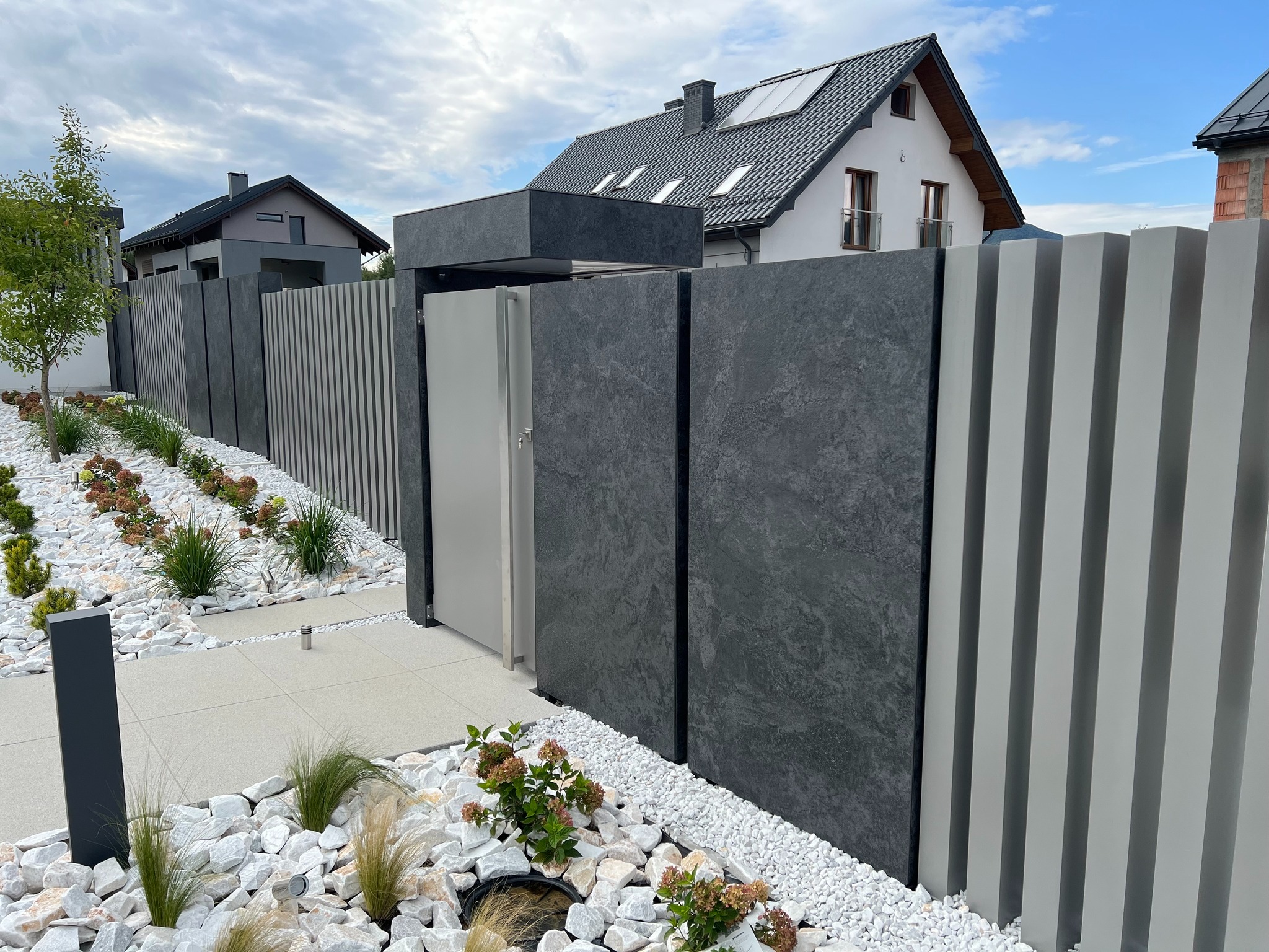 Nowoczesne ogrodzenie panelowe z elementami imitującymi kamień i furtką, otoczone białymi kamieniami i zielenią, na tle domu z panelami słonecznymi.