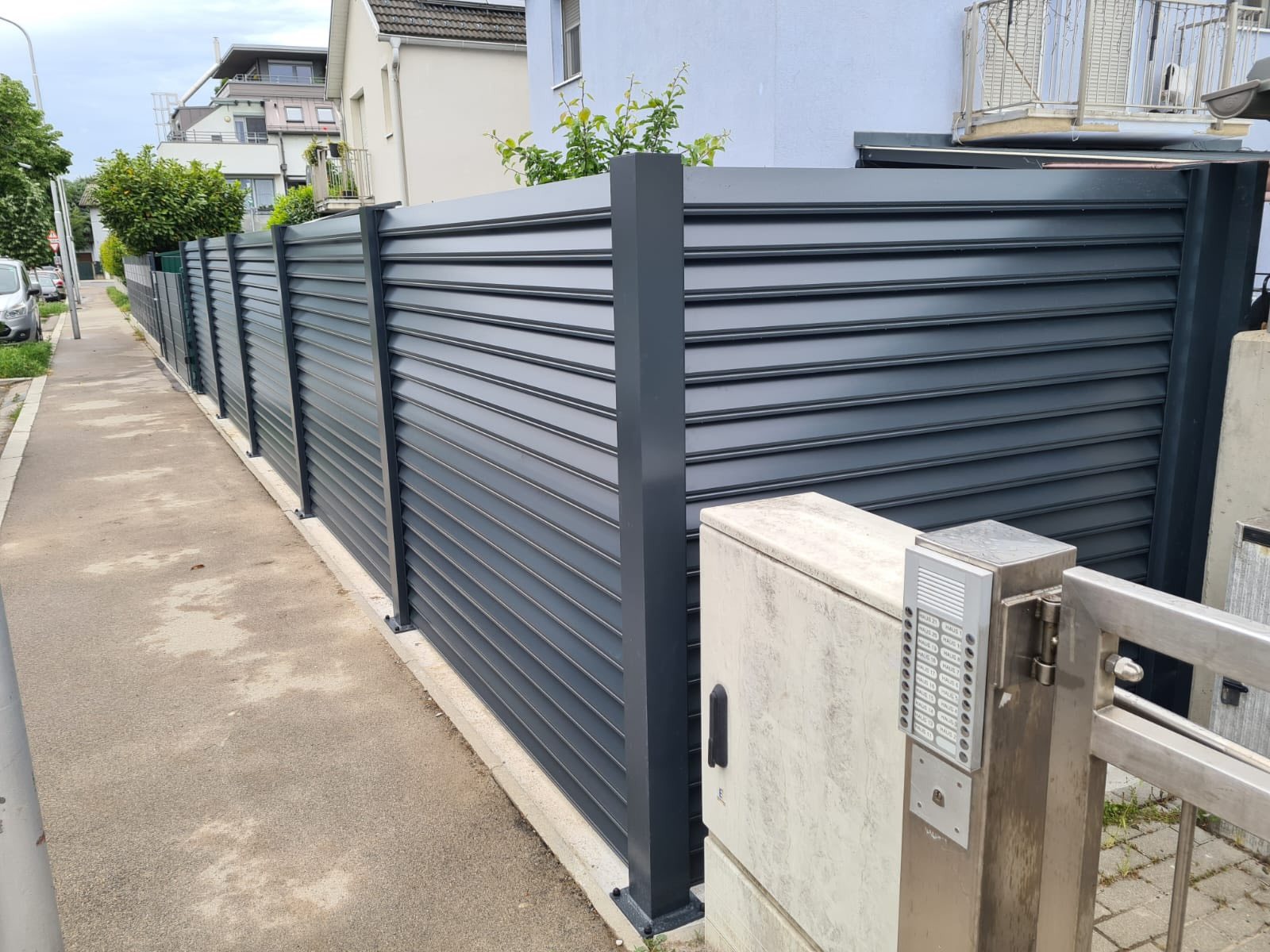 Nowoczesne, szare ogrodzenie lamelowe z metalu, widoczne od strony ulicy z domofonem przy furtce. Minimalistyczny design, poziome lamele, proste słupki.