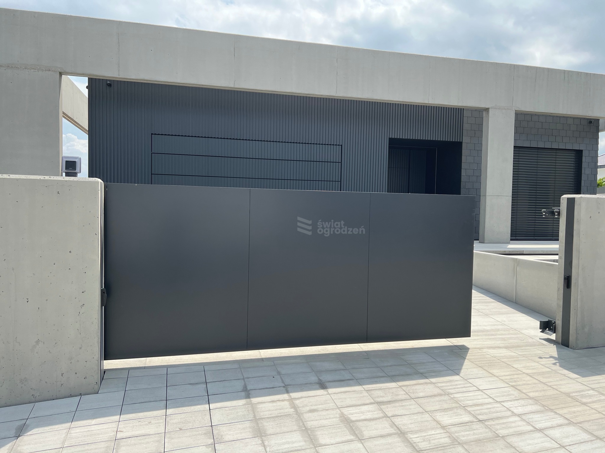 Nowoczesna, grafitowa brama przesuwna z logo w Orzechu, wkomponowana w minimalistyczną architekturę budynku. Betonowe słupki i jasna kostka brukowa.