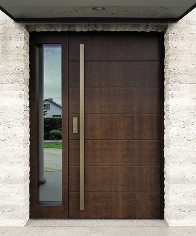 Nowoczesne, brązowe drzwi zewnętrzne z długim, pionowym uchwytem i bocznym przeszkleniem, osadzone w kamiennej ramie. Widoczny fragment domu w odbiciu szyby.