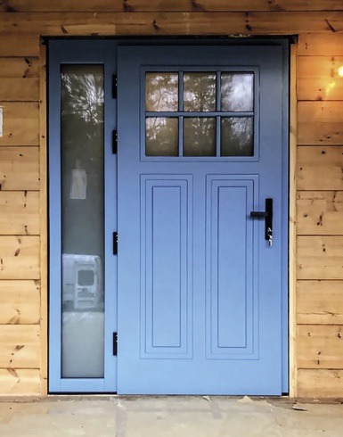 Błękitne drzwi zewnętrzne z szybą i wzorem kasetonowym, czarny uchwyt, drewniana elewacja budynku, delikatne światło, widok z bliska.