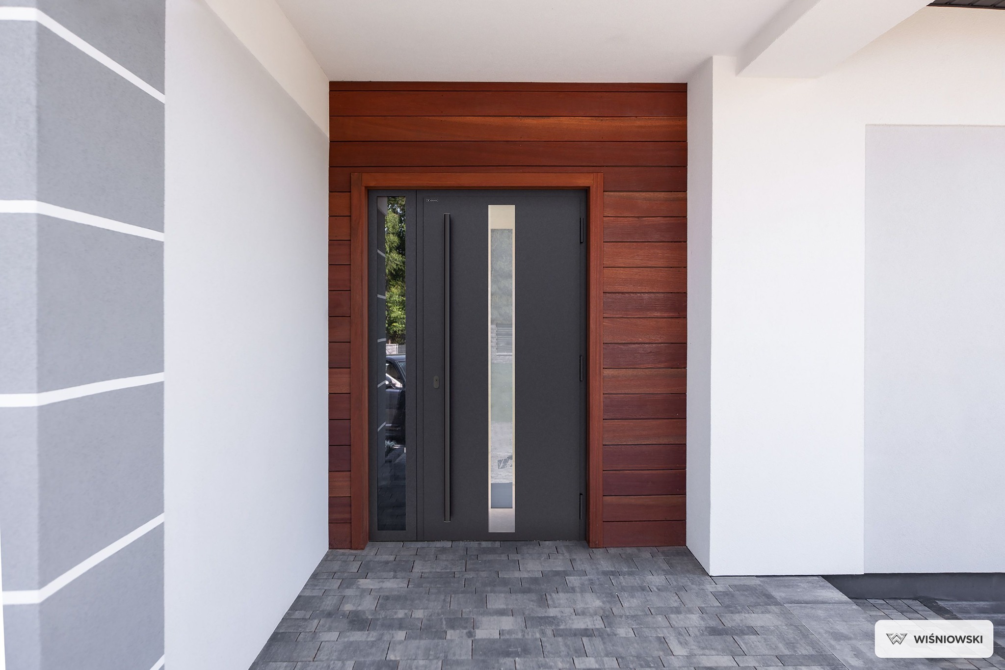 Szare drzwi zewnętrzne z szybą, osadzone w drewnianej obudowie, na tle białych ścian i szarej kostki brukowej. Minimalistyczny design, nowoczesne wykończenie.