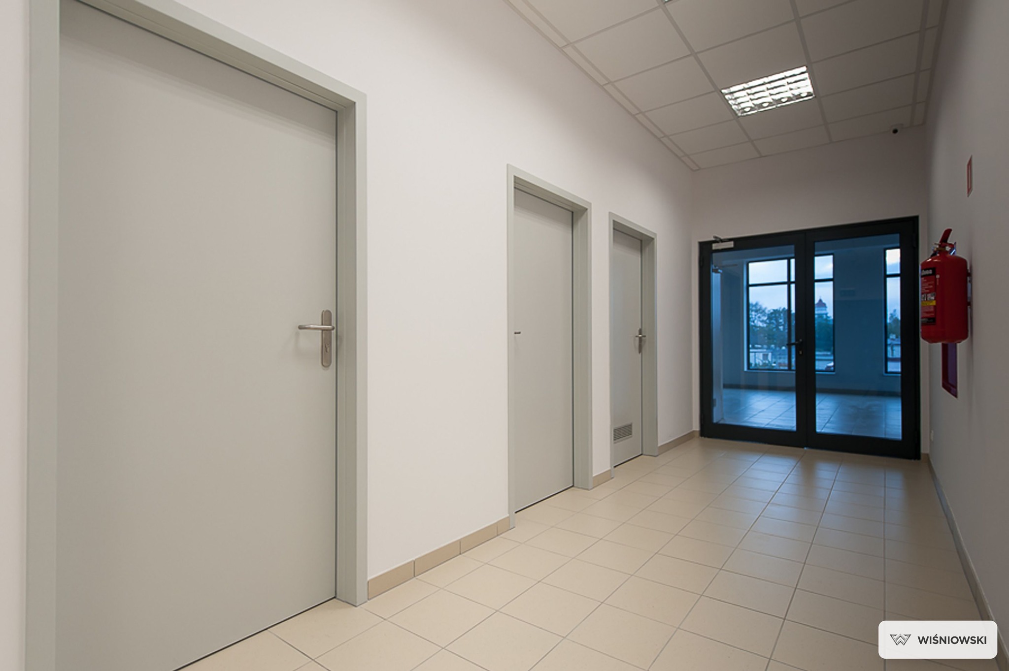 Korytarz z trzema parami drzwi w odcieniach szarości, z widocznymi klamkami i wentylacją. W tle dwuskrzydłowe drzwi szklane i gaśnica.
