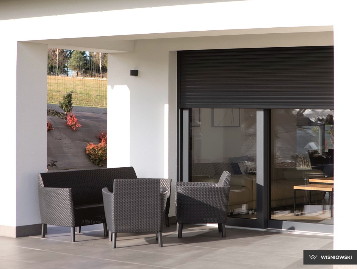 Zewnętrzna roleta w kolorze antracytu, zamontowana na dużym oknie przesuwnym. Widok na patio z meblami ogrodowymi i zielenią w tle. Nowoczesna architektura.